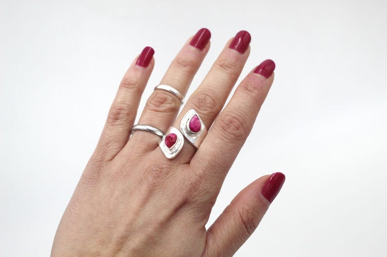 Może przedstawiać: Dłoń z trzema srebrnymi pierścionkami, w tym dwoma z dużymi, czerwonymi kamieniami w kształcie łezki. Pierścionki są na białym tle. Paznokcie pomalowane są na głęboki czerwony kolor.