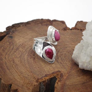 Może przedstawiać: Srebrny pierścionek z dwoma oprawami w kształcie łezki, z kt&oacute;rych każda zawiera żywy czerwony kamień szlachetny. Pierścionek jest prezentowany na teksturowanej drewnianej powierzchni, z białą formacją kryształową w tle. Biżuteria.
