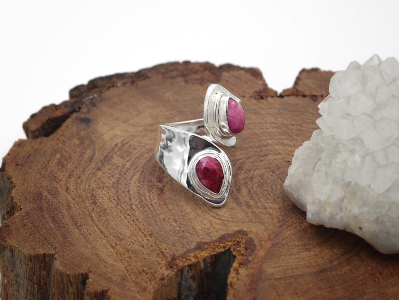 Może przedstawiać: Srebrny pierścionek z dwoma oprawami w kształcie łezki, z kt&oacute;rych każda zawiera żywy czerwony kamień szlachetny. Pierścionek jest prezentowany na teksturowanej drewnianej powierzchni, z białą formacją kryształową w tle. Biżuteria.