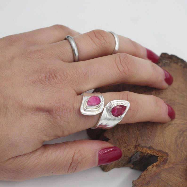 Może przedstawiać: Dłoń z trzema srebrnymi pierścionkami. Dwa pierścionki mają łezkowaty, jasno r&oacute;żowy kamień szlachetny osadzony w srebrnej oprawie. Trzeci pierścionek to prosta srebrna obrączka. Tło jest jasne z drewnianą powierzchnią.