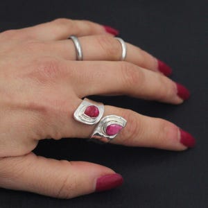 Może przedstawiać: Dwa srebrne pierścionki o nowoczesnym designie, każdy z żywym czerwonym kamieniem szlachetnym. Pierścionki są na dłoni z czerwonym lakierem do paznokci, na ciemnym tle. Kamienie szlachetne mają unikalny, organiczny kształt.