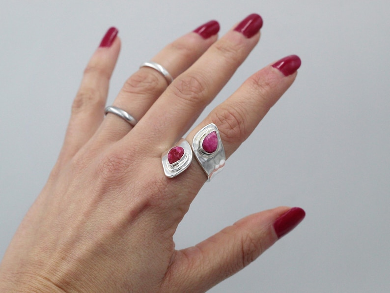 Może przedstawiać: Dłoń prezentuje trzy srebrne pierścionki, w tym dwa pierścionki z wyrazistymi, jaskrawoczerwonymi kamieniami szlachetnymi w kształcie łezki. Pierścionki mają młotkowaną fakturę. Paznokcie są pomalowane na pasujący czerwony kolor.