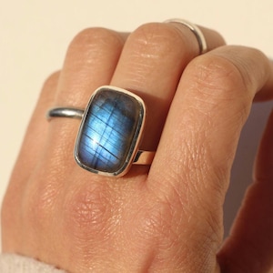 May include: A rectangular silver ring featuring a large, iridescent blue gemstone. The gemstone displays a captivating play of light, with flashes of blue and other colors. The ring is a statement piece, perfect for adding a touch of elegance.