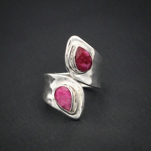 Könnte beinhalten: Ein silberner Ring mit zwei tropfenförmigen, tiefroten Edelsteinen. Der Ring hat ein Wickeldesign, bei dem die Edelsteine in einer Lünettenfassung gefasst sind. Der Ring wird vor einem dunklen Hintergrund gezeigt.