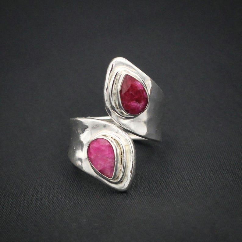 Może przedstawiać: Srebrny pierścionek z dwoma łzami w kształcie, głęboko czerwonymi kamieniami szlachetnymi. Pierścionek ma owijany design z kamieniami osadzonymi w oprawie ramowej. Pierścionek jest wyświetlany na ciemnym tle.