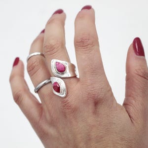 Może przedstawiać: Srebrny pierścionek z dwoma oprawami w kształcie łezki, z kt&oacute;rych każda zawiera żywy czerwony kamień szlachetny. Pierścionek owija się wok&oacute;ł palca, tworząc nowoczesny i elegancki design. Widoczny jest r&oacute;wnież prosty srebrny pierścionek.