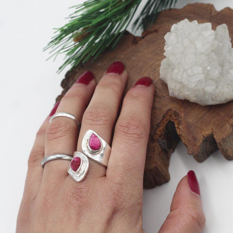 Może przedstawiać: Dłoń z trzema srebrnymi pierścionkami, w tym dwoma z żywymi r&oacute;żowymi kamieniami w kształcie łezki. Pierścionki są osadzone na tle naturalnego drewna z gałązką zieleni i białym skupiskiem kryształ&oacute;w.