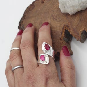 Może przedstawiać: Srebrny pierścionek z dwoma r&oacute;żowymi kamieniami szlachetnymi. Pierścionek owija się wok&oacute;ł palca, tworząc otwarty wz&oacute;r. Kamienie w kształcie łezki osadzone są w oprawie. Widoczny jest r&oacute;wnież prosty srebrny pierścionek.