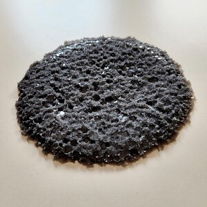 Puede incluir: Objeto circular poroso de color gris oscuro con una superficie texturizada. El objeto tiene apariencia de esponja con numerosos agujeros pequeños. La forma general es irregular, con un borde ligeramente áspero.
