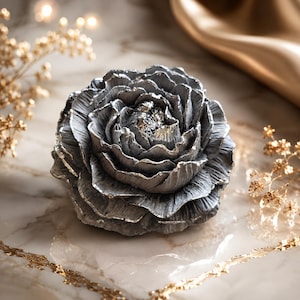 Könnte beinhalten: Ein skulpturales Blumenornament mit geschichteten, texturierten Blütenblättern in metallisch silberner Ausführung. Die Mitte der Blüte zeigt eine konzentrierte Ansammlung silberner Details, wobei die Form einer Pfingstrose ähnelt.