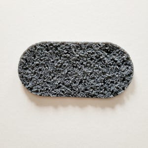 Könnte beinhalten: Ein grauer, oval geformter Bimsstein mit einer rauen, porösen Textur. Der Stein hat eine einfarbige Farbe und scheint aus kleinen, unregelmäßigen Teilen zu bestehen. Der Bimsstein wird zum Peelen der Haut verwendet.