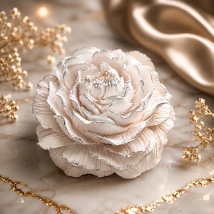 Könnte beinhalten: Ein skulpturales Blumenornament mit geschichteten, cremeweißen Blütenblättern und metallisch silbernen Rändern. Die Blume hat eine dichte, abgerundete Form mit komplexen, gerüschten Blütenblatttexturen, die ein geschichtetes Erscheinungsbild erzeugen.