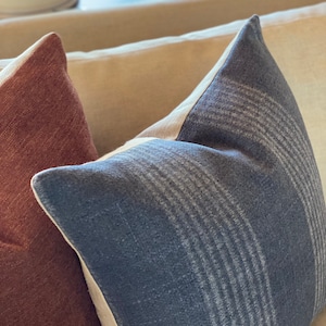May include: Close-up of three decorative throw pillows. The front pillow is a dark blue with a striped pattern. The pillow to the left is a reddish-brown color. The pillow in the background is a light beige color.