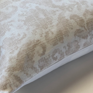 May include: A beige throw pillow with a subtle, light brown patterned design. The pillow has a white border around the edges.