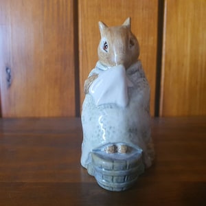 May include: A porcelain figurine of a brown and white rabbit wearing a blue robe and holding a white cloth. The rabbit is sitting on a wooden bucket.