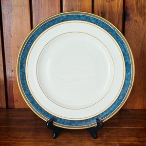 Peut inclure: Une assiette en c&eacute;ramique blanche avec un bord dor&eacute; et une bordure d&eacute;corative bleue. L'assiette est pr&eacute;sent&eacute;e sur un support noir sur fond de bois. L'assiette a un design classique et &eacute;l&eacute;gant.