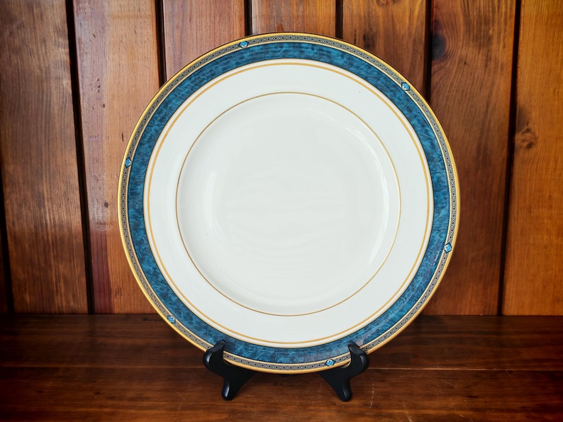 Peut inclure: Une assiette en c&eacute;ramique blanche avec un bord dor&eacute; et une bordure d&eacute;corative bleue. L'assiette est pr&eacute;sent&eacute;e sur un support noir sur fond de bois. L'assiette a un design classique et &eacute;l&eacute;gant.