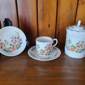 May include: A set of white porcelain teacups and saucers with a floral pattern featuring butterflies and roses. The set includes a small bowl, a teacup and saucer, and a lidded jar.
