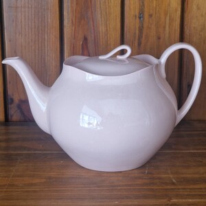 May include: A pale pink ceramic teapot with a curved spout, a loop handle, and a lid with a curled handle. The teapot has a rounded body and sits on a wooden surface. The background is a wooden wall.