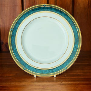 Peut inclure: Une assiette en porcelaine blanche avec une bordure turquoise et dor&eacute;e. L'assiette pr&eacute;sente une bande turquoise avec un motif g&eacute;om&eacute;trique dor&eacute; et une bordure dor&eacute;e. L'assiette est pr&eacute;sent&eacute;e sur un support blanc.