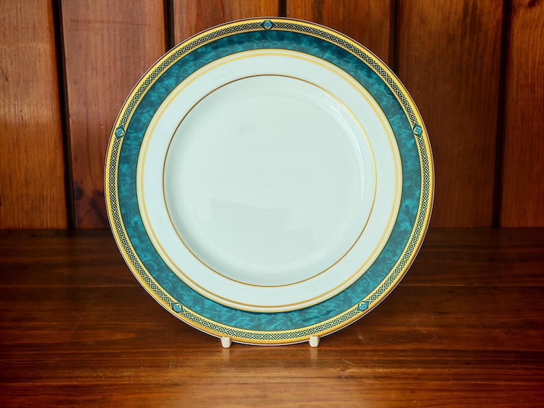Peut inclure: Une assiette en porcelaine blanche avec une bordure turquoise et dor&eacute;e. L'assiette pr&eacute;sente une bande turquoise avec un motif g&eacute;om&eacute;trique dor&eacute; et une bordure dor&eacute;e. L'assiette est pr&eacute;sent&eacute;e sur un support blanc.