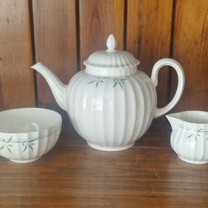 May include: A white ceramic tea set comprising a teapot, creamer jug, and bowl. The teapot has a lid and a curved spout. The set features a ribbed design with blue floral accents. The set is on a wooden surface.