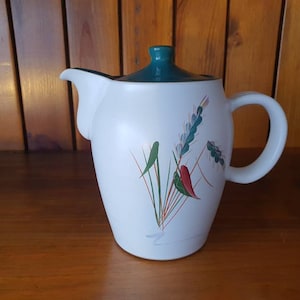 May include: White ceramic teapot with a green lid and a floral design featuring green, red, and white leaves. The teapot has a handle on the side.