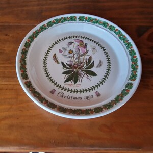 May include: A white ceramic Christmas plate with a green holly border. The center features a botanical illustration of a Helleborus Niger, or Christmas Rose, with the text "Christmas 1993".