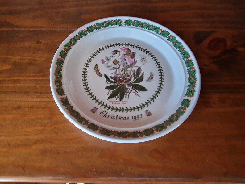 May include: A white ceramic Christmas plate with a green holly border. The center features a botanical illustration of a Helleborus Niger, or Christmas Rose, with the text "Christmas 1993".