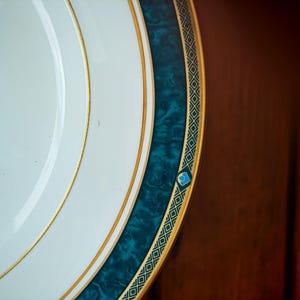 Peut inclure: Gros plan sur une assiette blanche avec une bordure turquoise et dor&eacute;e. L'assiette a un bord dor&eacute; et une bande turquoise avec un effet marbr&eacute;. La bande turquoise est bord&eacute;e d'une garniture dor&eacute;e avec un motif g&eacute;om&eacute;trique.