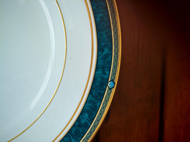 Peut inclure: Gros plan sur une assiette blanche avec une bordure turquoise et dor&eacute;e. L'assiette a un bord dor&eacute; et une bande turquoise avec un effet marbr&eacute;. La bande turquoise est bord&eacute;e d'une garniture dor&eacute;e avec un motif g&eacute;om&eacute;trique.