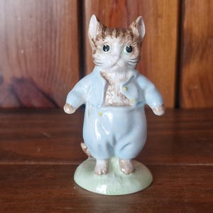 Puede incluir: Figura de cerámica de un gato con un atuendo azul claro. El gato tiene pelaje marrón y canela y ojos verdes. La figura está sobre una pequeña base ovalada verde. El atuendo incluye una chaqueta y pantalones.