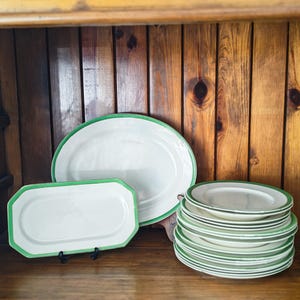 May include: A collection of white ceramic serving dishes with green trim. The set includes oval platters and rectangular dishes, along with a stack of smaller plates. The dishes are displayed on a wooden shelf against a wooden background.