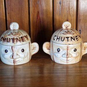 May include: Two ceramic chutney jars with a whimsical face design. Each jar has a brown lid with the word "CHUTNEY" written in brown lettering.