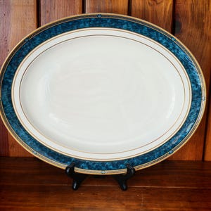 Peut inclure: Un plat de service ovale avec un centre blanc et une bordure d&eacute;corative. La bordure pr&eacute;sente un motif bleu canard fonc&eacute; avec des accents dor&eacute;s. Le plat est pr&eacute;sent&eacute; sur un support noir sur un fond en bois. Une pi&egrave;ce classique pour servir des aliments.