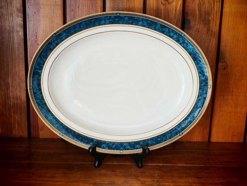 Peut inclure: Un plat de service ovale avec un centre blanc et une bordure d&eacute;corative. La bordure pr&eacute;sente un motif bleu canard fonc&eacute; avec des accents dor&eacute;s. Le plat est pr&eacute;sent&eacute; sur un support noir sur un fond en bois. Une pi&egrave;ce classique pour servir des aliments.