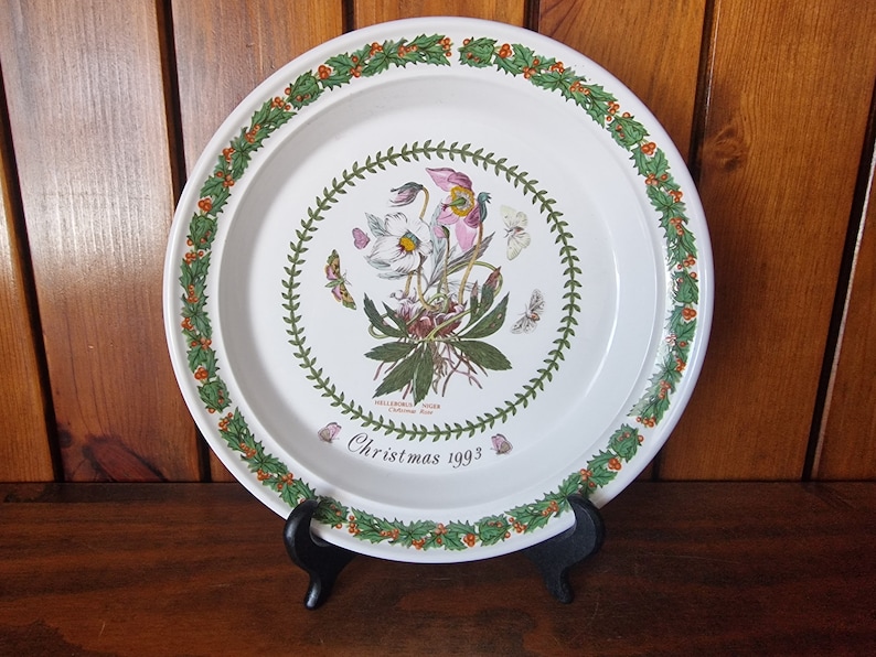 May include: White ceramic Christmas plate with a green holly border and a floral design in the center. The plate features a white flower with pink accents and green leaves. The text "Christmas 1993" is printed on the plate.