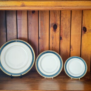 Peut inclure: Trois assiettes en c&eacute;ramique blanche avec une bordure bleue et dor&eacute;e, expos&eacute;es sur une &eacute;tag&egrave;re en bois. Les assiettes sont de tailles diff&eacute;rentes, la plus grande &eacute;tant &agrave; l'avant. L'arri&egrave;re-plan est une armoire en bois.