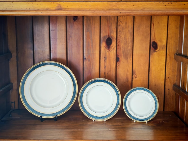 Peut inclure: Trois assiettes en c&eacute;ramique blanche avec une bordure bleue et dor&eacute;e, expos&eacute;es sur une &eacute;tag&egrave;re en bois. Les assiettes sont de tailles diff&eacute;rentes, la plus grande &eacute;tant &agrave; l'avant. L'arri&egrave;re-plan est une armoire en bois.