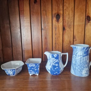 May include: A collection of blue and white ceramic serving pieces, including a rectangular dish, a small square planter, a pitcher, and a larger water jug. The pieces feature a floral pattern and are arranged on a wooden surface against a wooden backdrop.