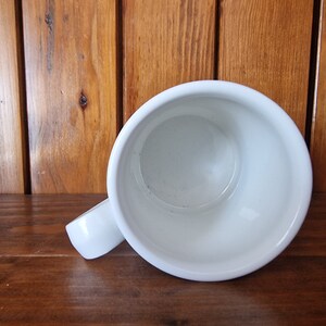 May include: White ceramic mug with a handle. The mug is empty and is sitting on a wooden surface.