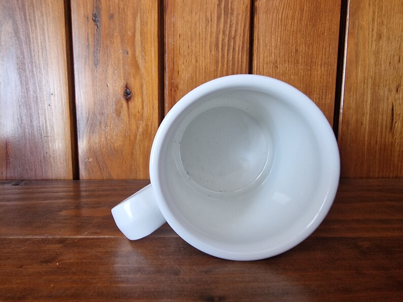 May include: White ceramic mug with a handle. The mug is empty and is sitting on a wooden surface.