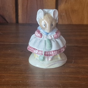 May include: A ceramic figurine of a mouse wearing a white dress with a red and pink trim, a blue scarf, and a white bonnet. The mouse is standing on a small, round base. The figurine has a vintage aesthetic.