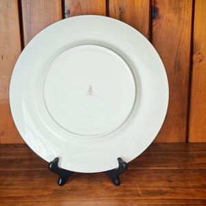 Peut inclure: Une grande assiette en c&eacute;ramique blanc cass&eacute; avec un large bord, pr&eacute;sent&eacute;e sur un support noir. Le dessous de l'assiette est visible, r&eacute;v&eacute;lant une zone centrale en retrait et un petit embl&egrave;me dor&eacute;. L'arri&egrave;re-plan est une surface en bois.