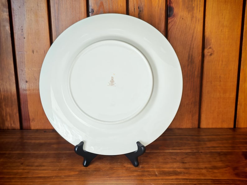 Peut inclure: Une grande assiette en c&eacute;ramique blanc cass&eacute; avec un large bord, pr&eacute;sent&eacute;e sur un support noir. Le dessous de l'assiette est visible, r&eacute;v&eacute;lant une zone centrale en retrait et un petit embl&egrave;me dor&eacute;. L'arri&egrave;re-plan est une surface en bois.