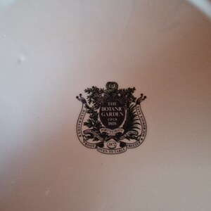 May include: A white ceramic plate with a black and green crest featuring the text "The Botanic Garden circa 1818" and "Dishwasher Proof Oven to Table Freezer", surrounded by a wreath of leaves.