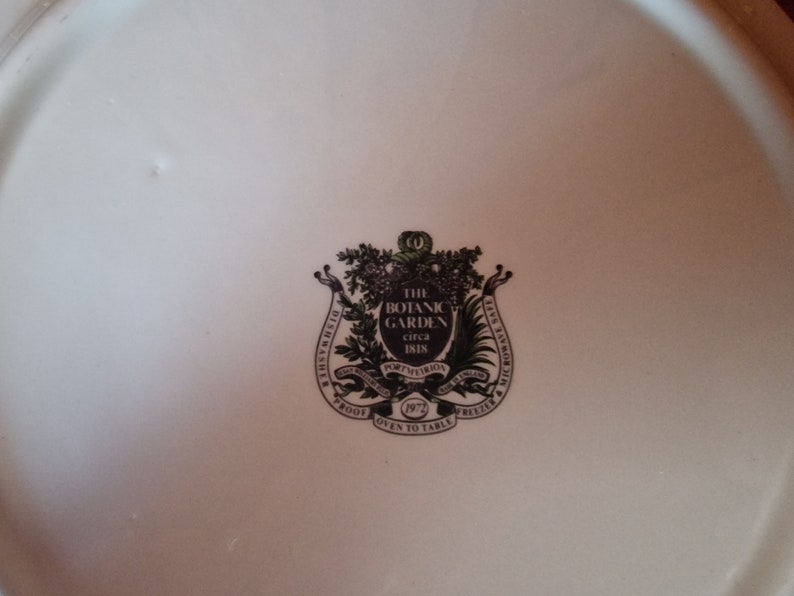 May include: A white ceramic plate with a black and green crest featuring the text "The Botanic Garden circa 1818" and "Dishwasher Proof Oven to Table Freezer", surrounded by a wreath of leaves.