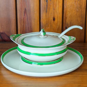 May include: A vintage cream soup tureen with a lid and spoon, presented on a round plate. The tureen and plate are decorated with bright green and gold trim. The lid has a decorative finial.