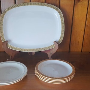 May include: A set of white ceramic plates with a gold rim. The set includes a large oval serving platter and four smaller round plates.