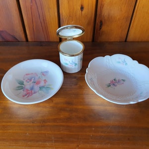 Könnte beinhalten: Ein Set aus drei weißen Porzellantellern mit Blumendesigns. Die Teller zeigen ein zartes Blumenmuster mit rosa, blauen und grünen Akzenten. Das Set enthält einen kleinen runden Teller, einen größeren runden Teller und einen kleinen runden Teller mit Deckel.